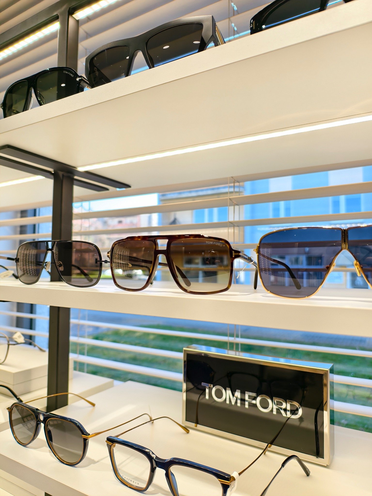 Różnorodne okulary korekcyjne i przeciwsłoneczne marki Tom Ford, eksponowane na półkach w salonie optycznym, z żaluzjami w tle.