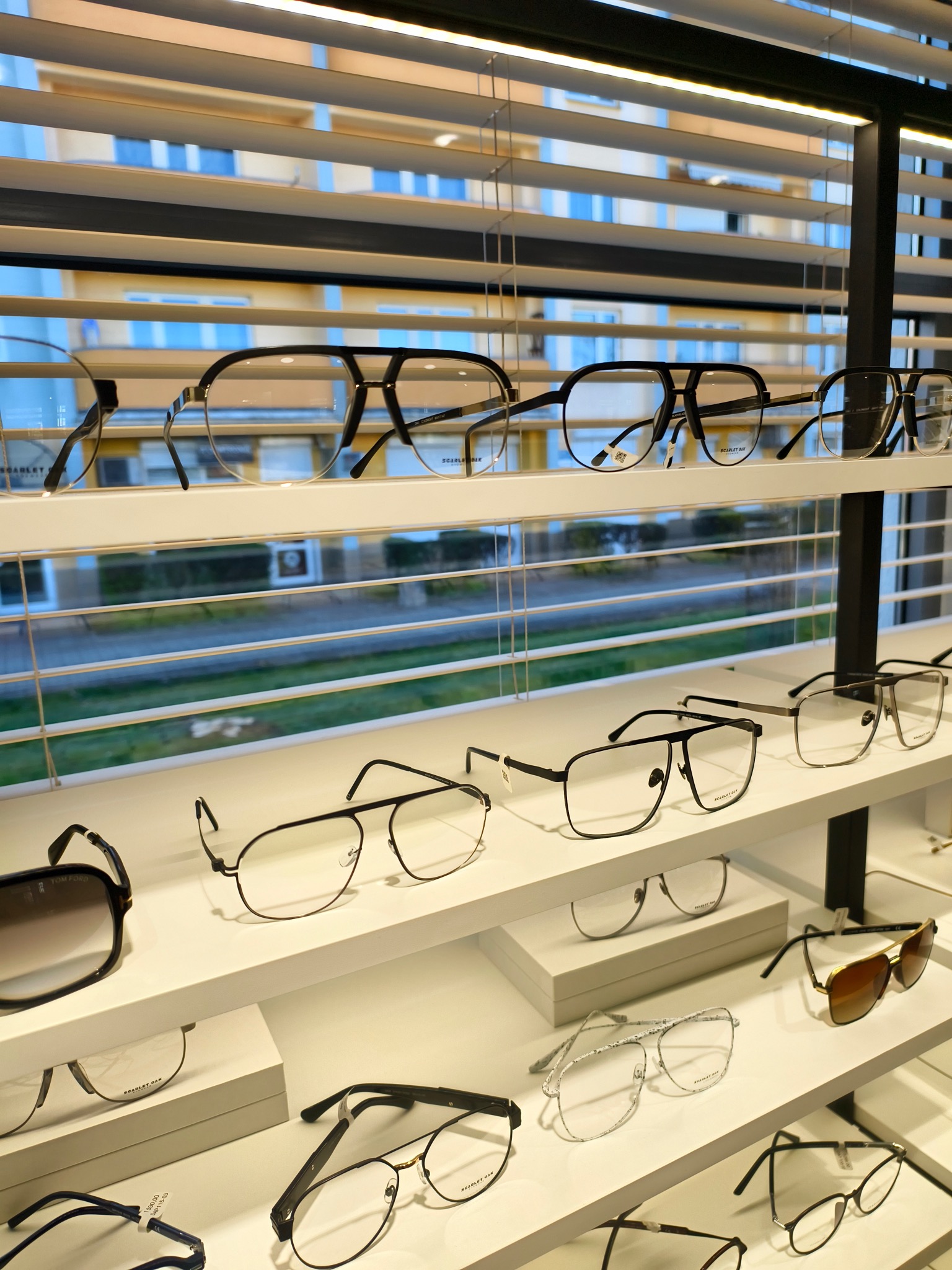 Różnorodne oprawki okularów korekcyjnych i przeciwsłonecznych na białych półkach w salonie optycznym, widoczne przez żaluzje okienne.