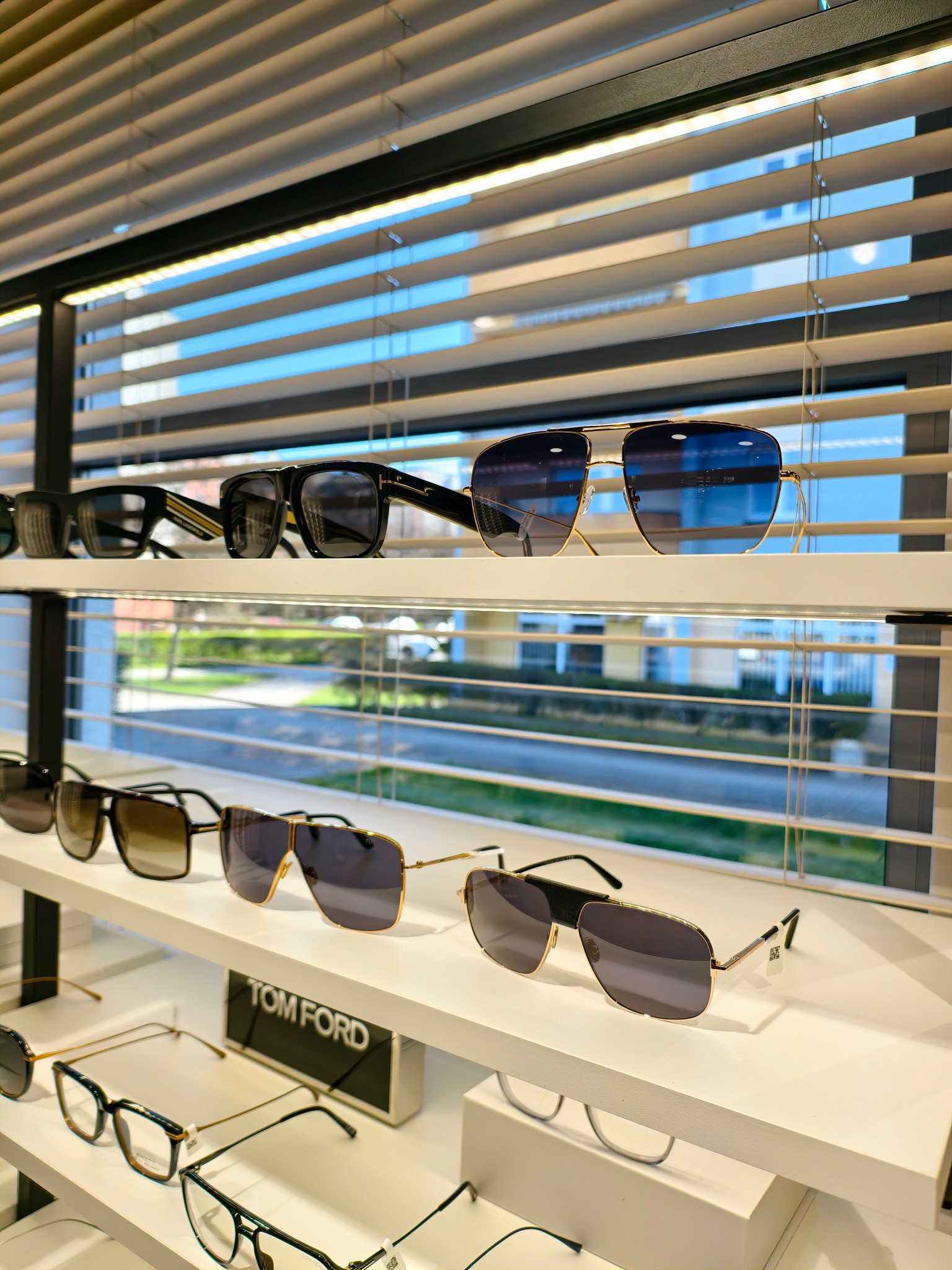 Ekspozycja okularów korekcyjnych i przeciwsłonecznych różnych marek na białych półkach sklepowych, z widokiem na budynki przez żaluzje w tle.