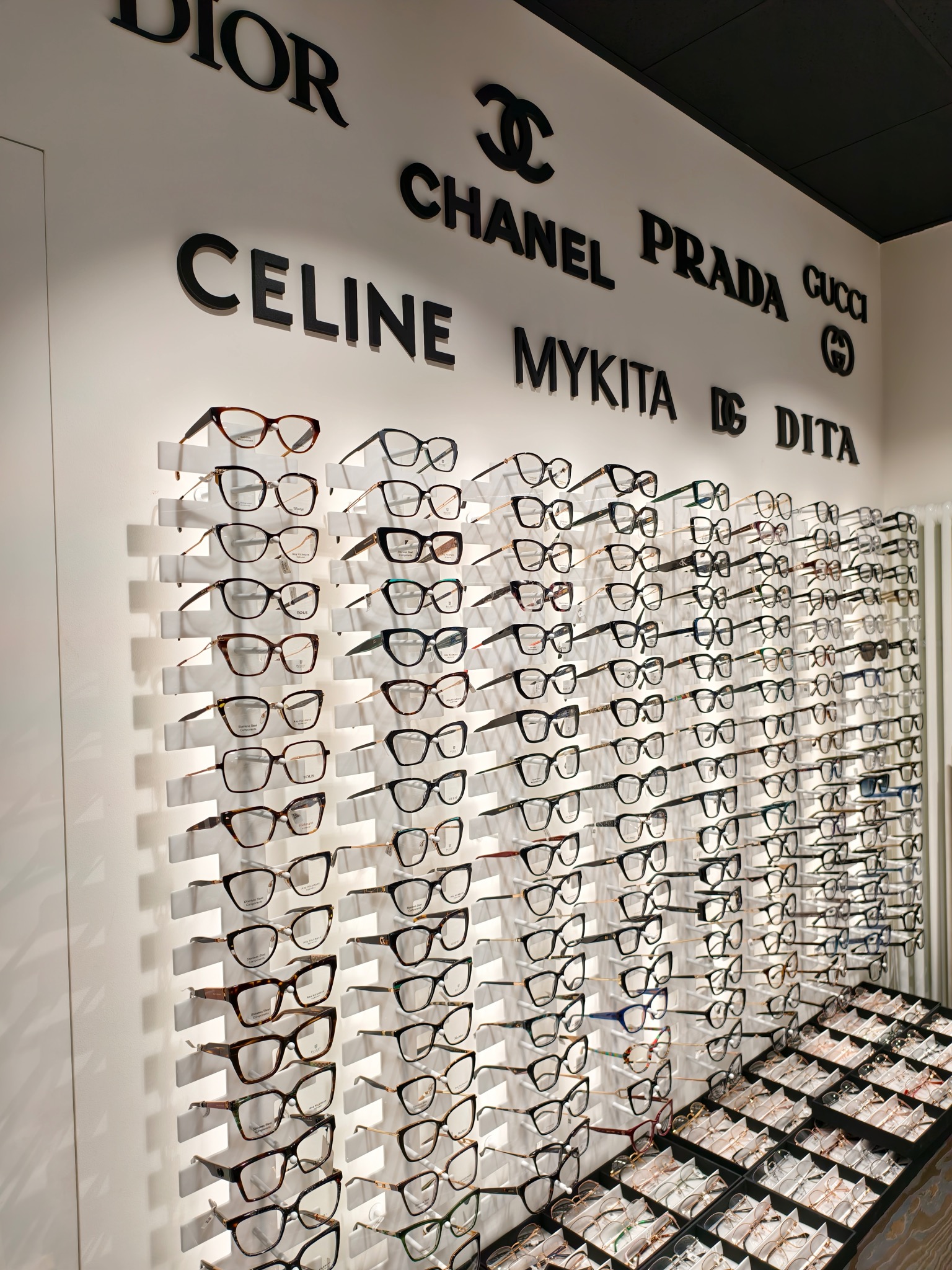 Ekspozycja oprawek okularów w salonie optycznym, marki takie jak Dior, Chanel, Prada, Celine, Mykita i Dita na ścianie, różnorodne modele w rzędach i w gablotach.