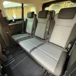 Wnętrze samochodu z trzema rzędami siedzeń, szara tapicerka z geometrycznym wzorem, czarne gumowe dywaniki na podłodze, butelka napoju w bocznej kieszeni.