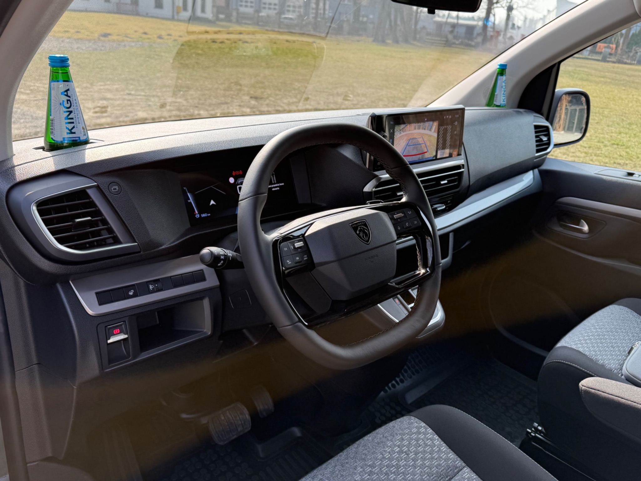 Wnętrze nowoczesnego samochodu dostawczego Peugeot z widocznym kokpitem, kierownicą, ekranem nawigacji z włączoną kamerą cofania oraz butelkami wody mineralnej Kinga umieszczonymi na desce...