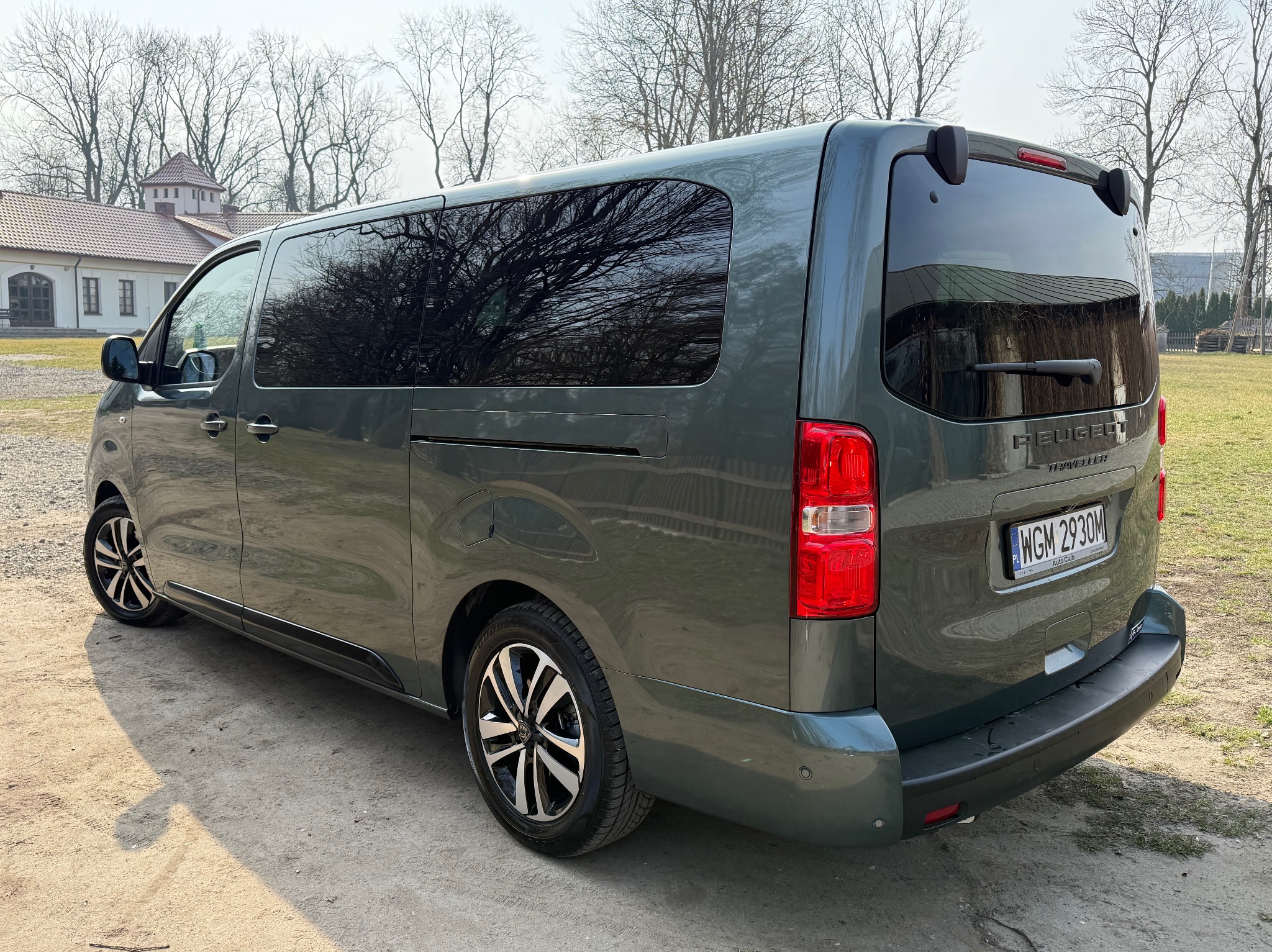 Szary bus Peugeot Traveller zaparkowany w Warszawie na żwirowym podłożu, z widocznym budynkiem w tle i odbiciem drzew w oknach.