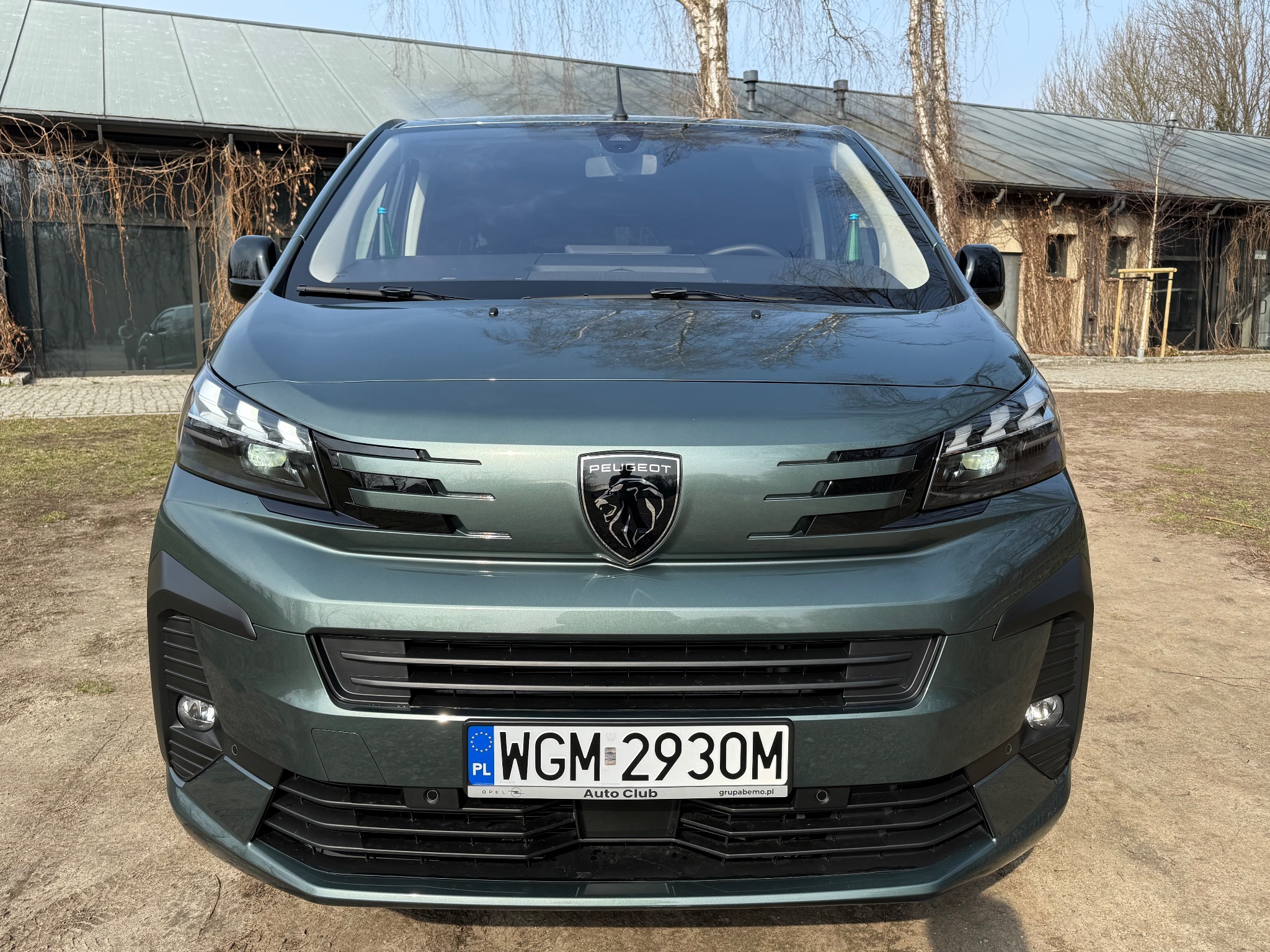 Przód oliwkowozielonego Peugeota z widoczną tablicą rejestracyjną WGM 2930M na tle budynku z szarą blachą i pnączami.