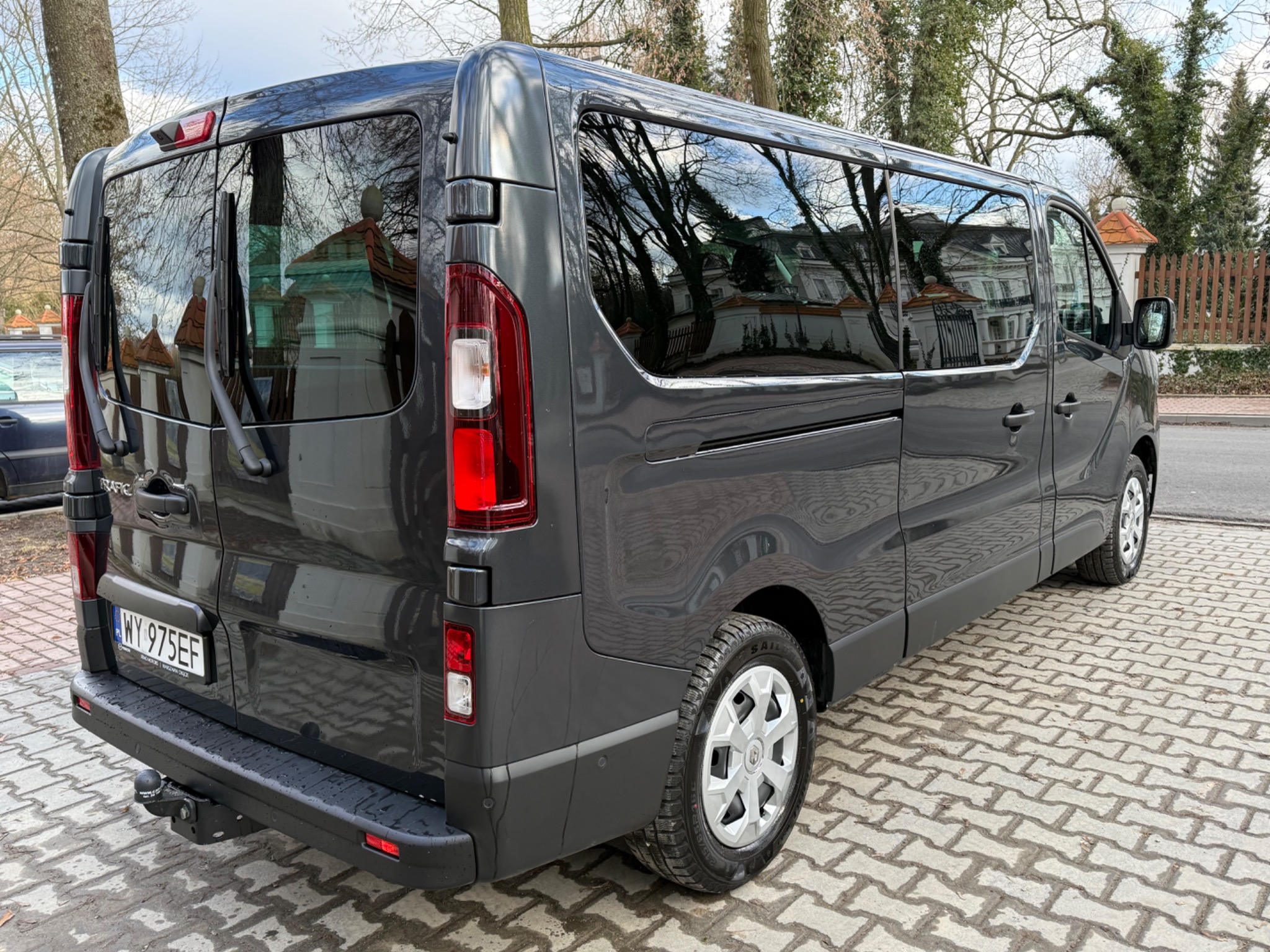 Szary bus marki Renault Trafic, widok z tyłu, z widocznymi detalami jak hak holowniczy i tablica rejestracyjna WY 975EF, zaparkowany na brukowanej nawierzchni z odbiciem budynków w oknach.