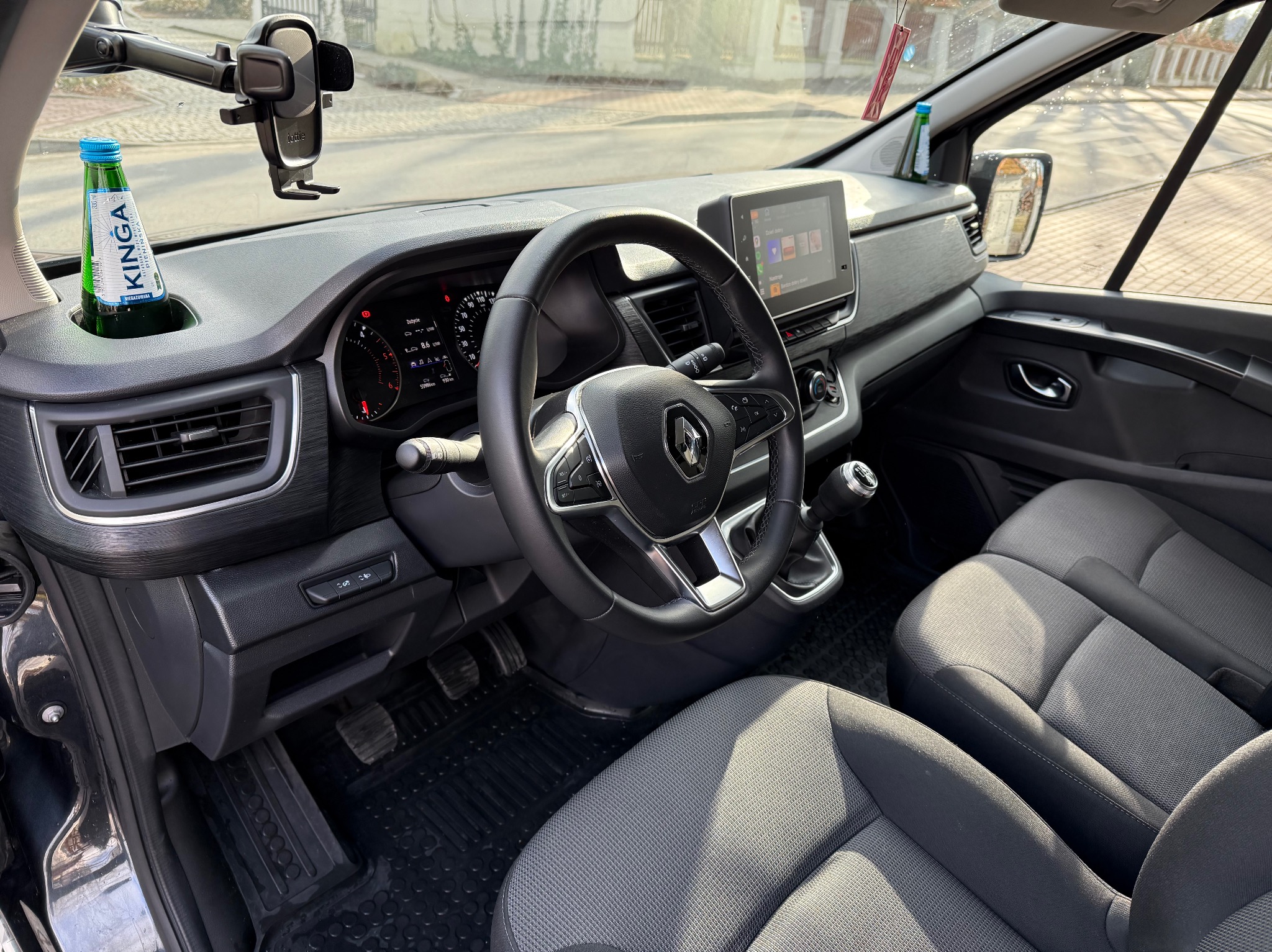 Wnętrze Renault Trafic z widoczną butelką wody Kinga w uchwycie, uchwytem na telefon i szarą tapicerką foteli.