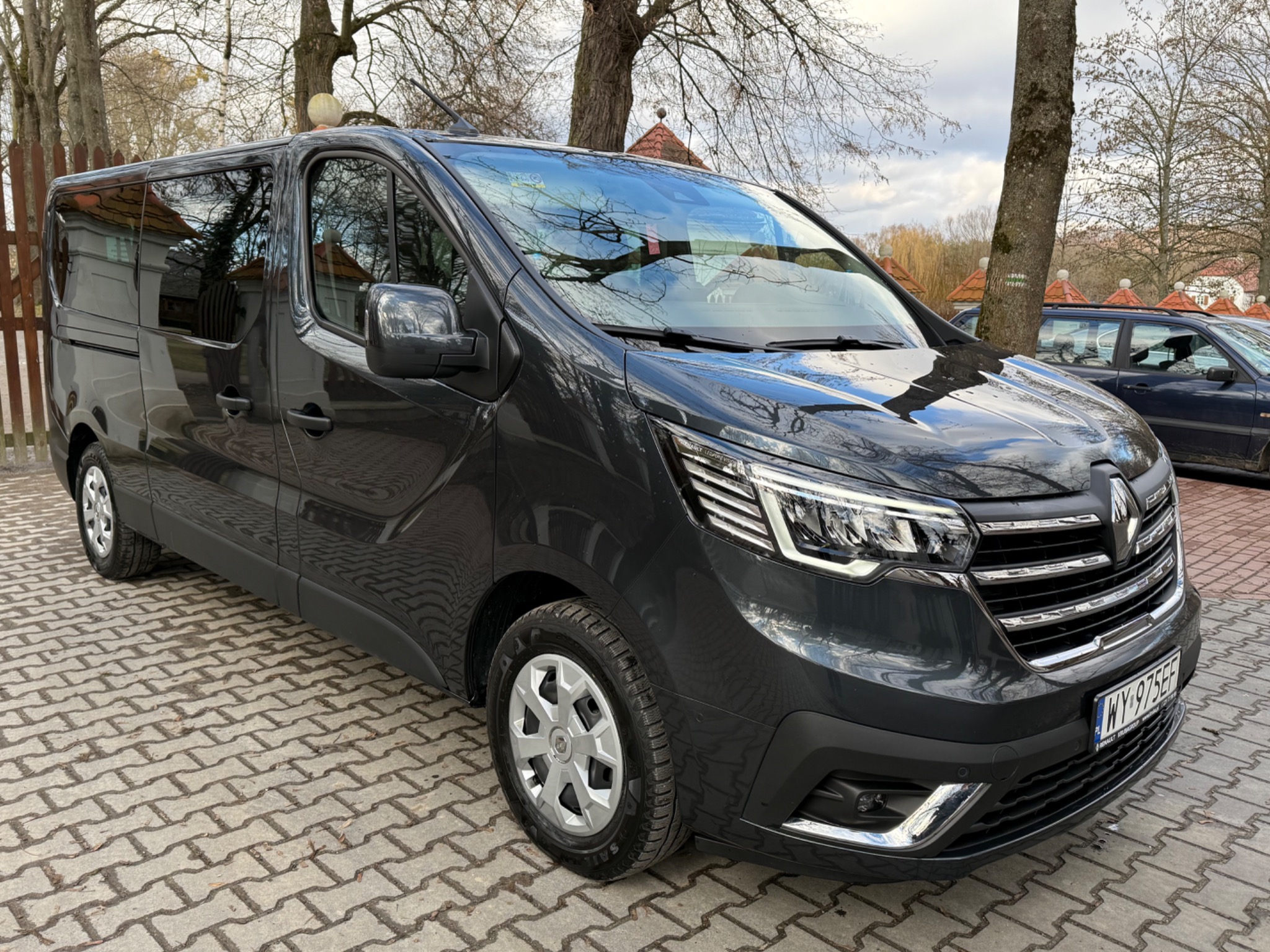 Ciemnoszary Renault Trafic, zaparkowany na brukowanej nawierzchni, z widocznym numerem rejestracyjnym i odbiciami w szybach.