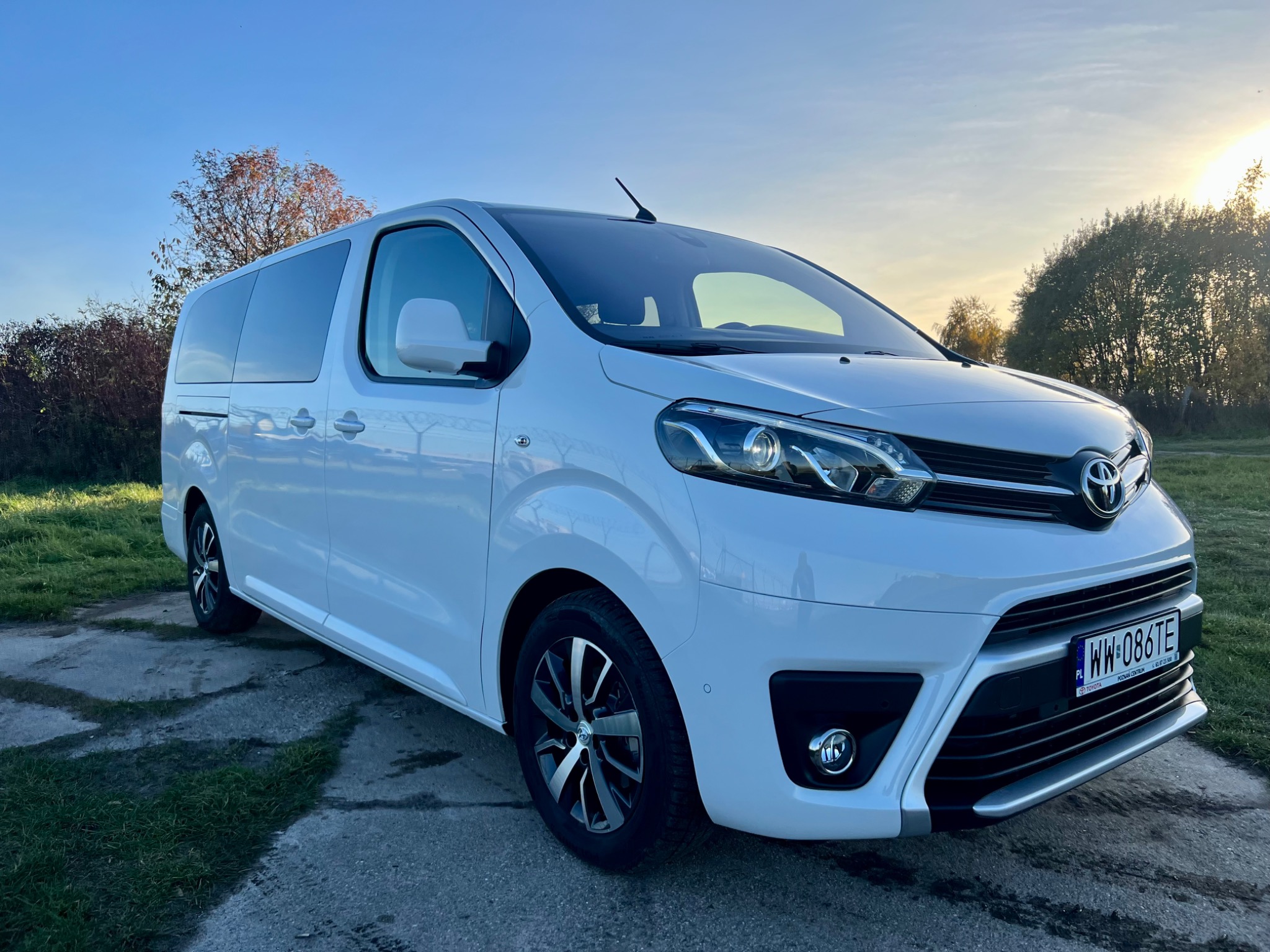Biały van Toyota Proace Verso z przyciemnianymi szybami, zaparkowany na betonowej powierzchni z zielonymi kępami trawy, sfotografowany pod słońce.