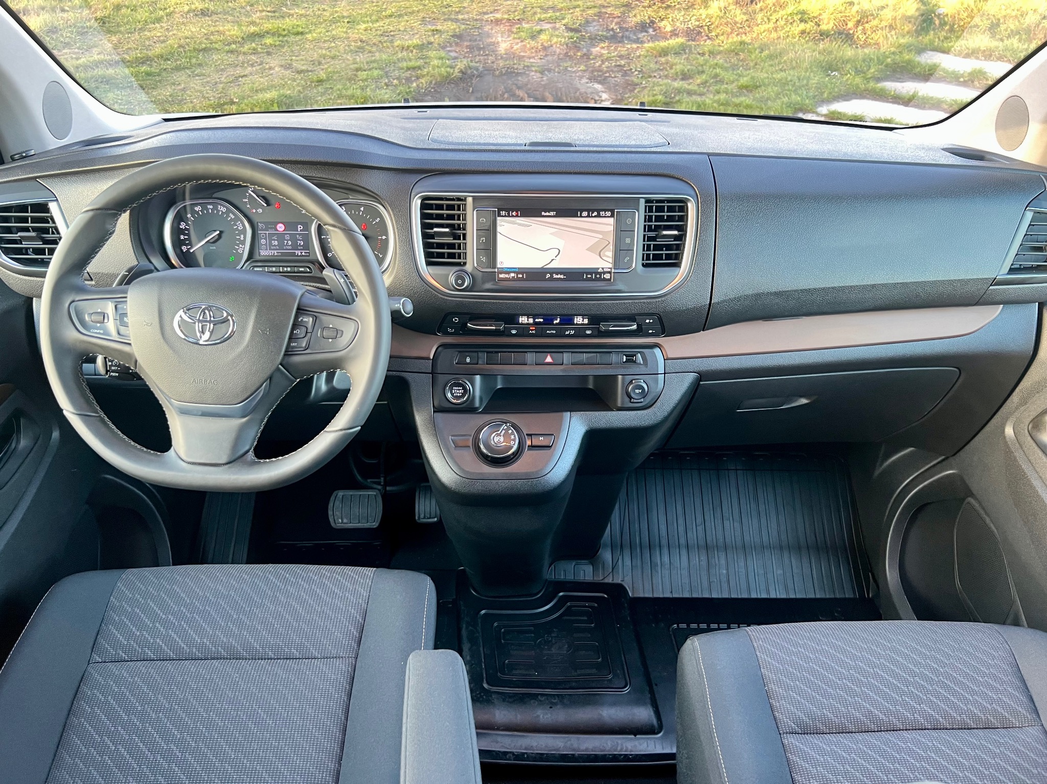 Wnętrze samochodu Toyota Proace Verso z widocznym kokpitem, kierownicą z logo, nawigacją na desce rozdzielczej i czarnymi dywanikami pod nogami.
