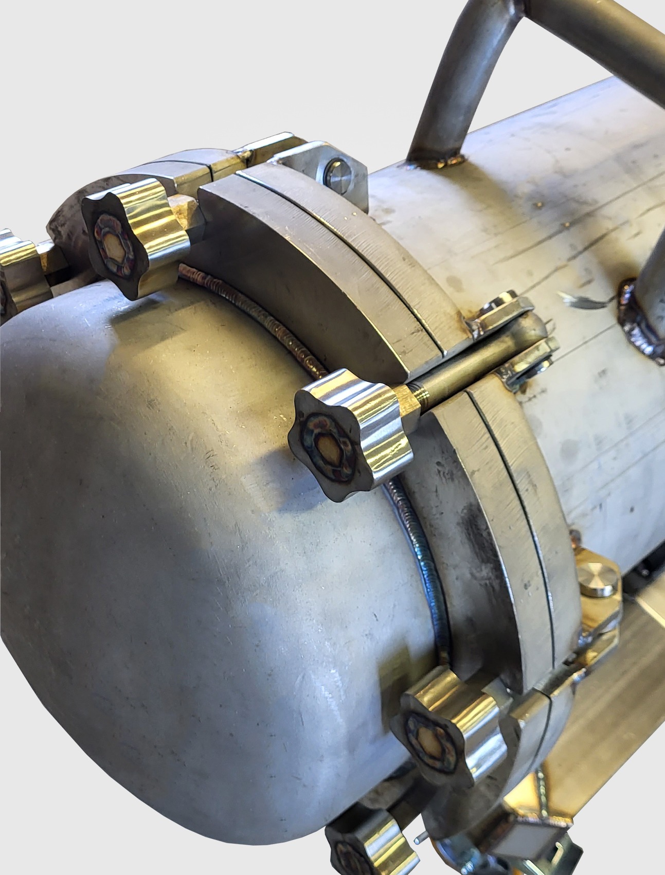 Szczegółowe ujęcie metalowego zbiornika z elementami mocującymi i uchwytem, widoczne ślady spawania, całość w odcieniach szarości i srebra, oświetlenie studyjne.