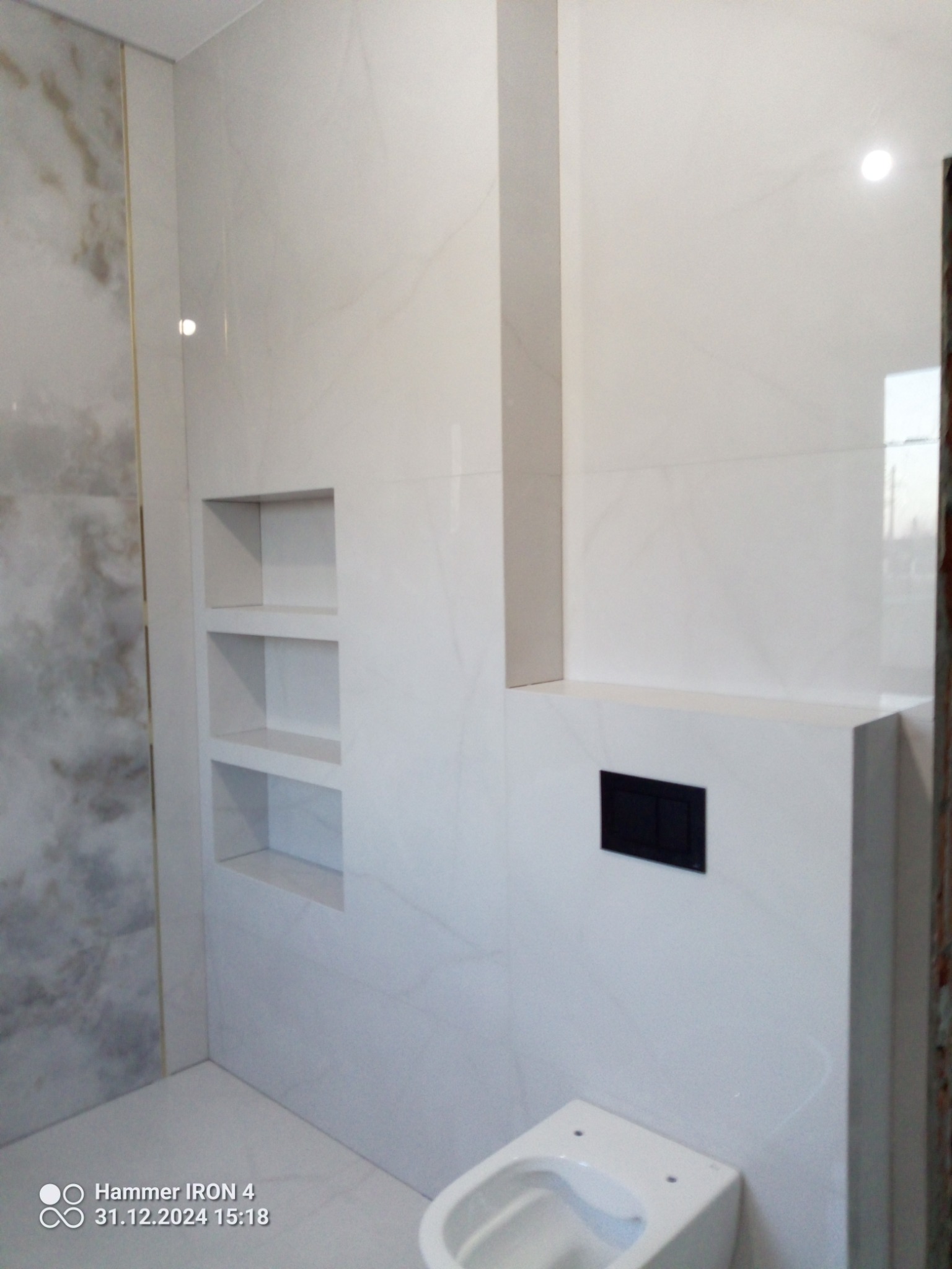 Biała łazienka z wykończeniem marmurowym, zabudową WC i wnękami półkowymi. Minimalistyczny design, płytki imitujące marmur, czarny przycisk spłukiwania.