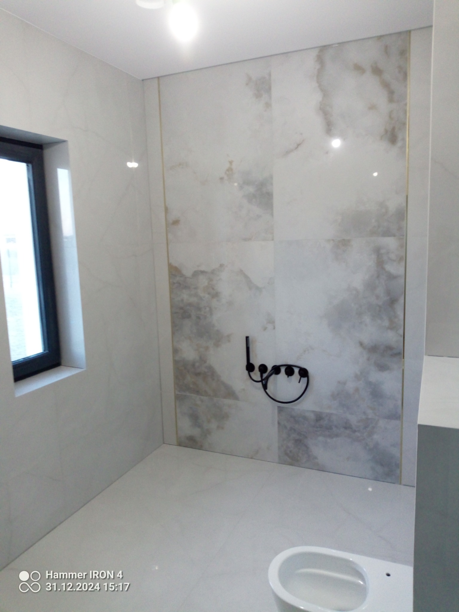 Wykończona łazienka w Brzezinach: białe płytki imitujące marmur na ścianach i podłodze, czarna armatura i okno. Minimalistyczny design, eleganckie wykończenie.