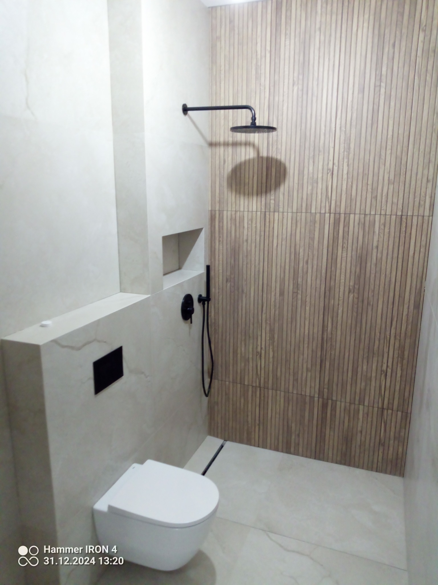 Nowoczesna łazienka z podwieszaną toaletą, deszczownicą i ścianą wykończoną drewnopodobnymi panelami. Minimalistyczny design i jasne płytki podłogowe.