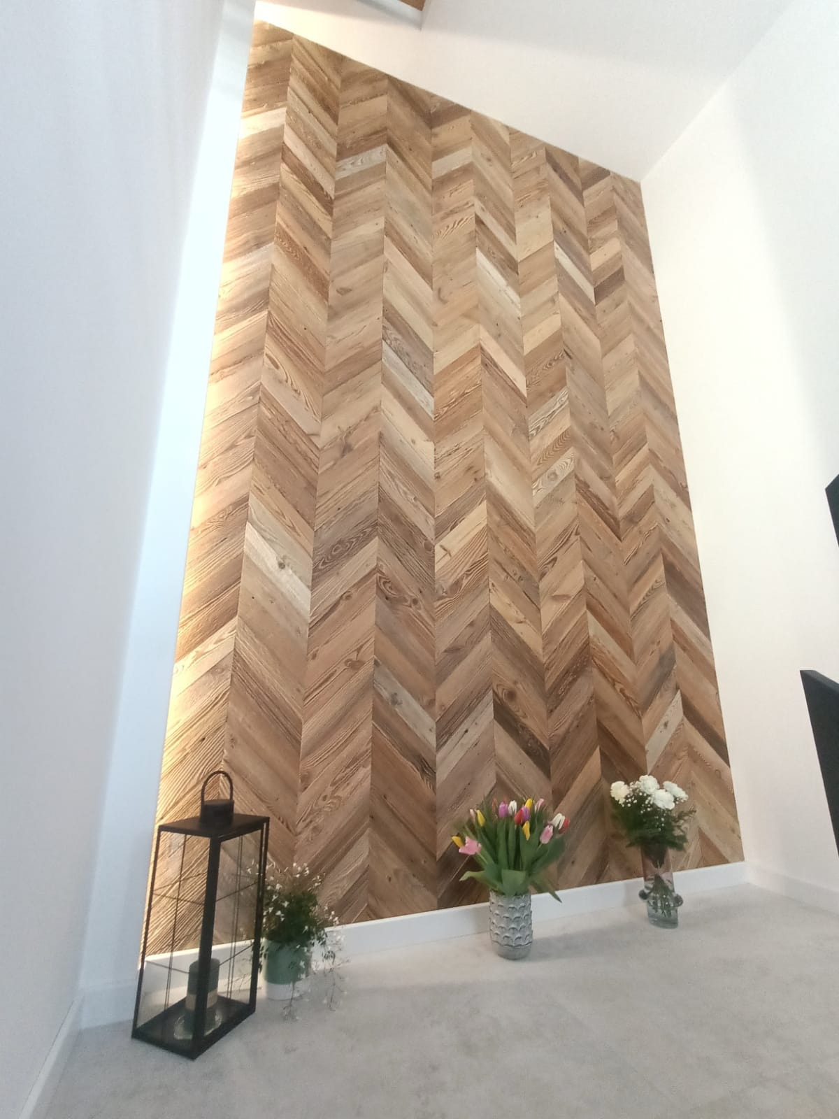 Ściana wykończona drewnianą mozaiką w jodełkę, oświetlona z lewej strony. Dekoracje: lampion, bukiety kwiatów. Jasne wnętrze, minimalistyczny styl.