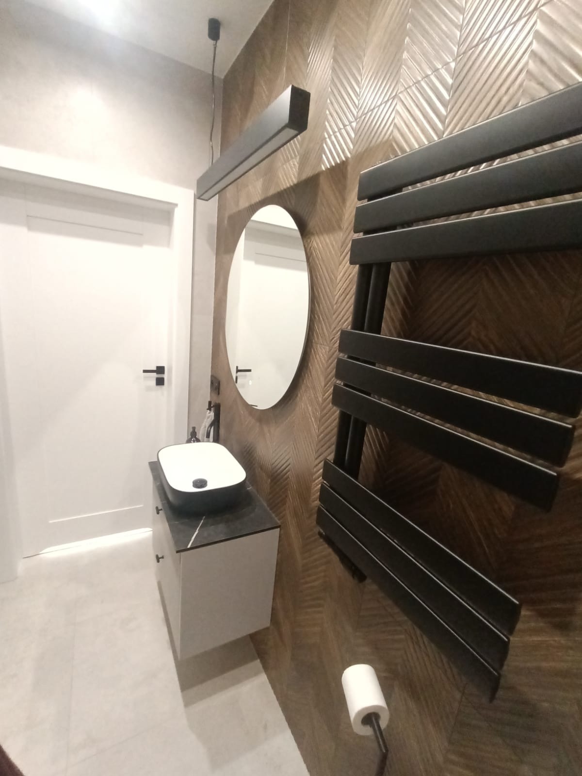 Nowoczesna łazienka z umywalką nablatową, lustrem i grzejnikiem drabinkowym na ścianie wyłożonej płytkami w geometryczny wzór. Minimalistyczne oświetlenie.