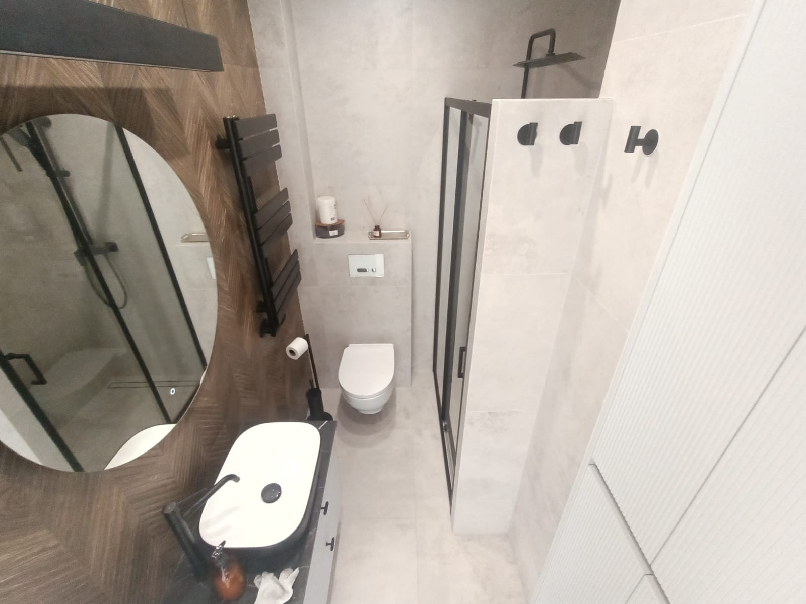 Nowoczesna łazienka z owalnym lustrem, czarną armaturą i kabiną prysznicową typu walk-in. Drewnopodobne płytki i minimalistyczne dodatki.