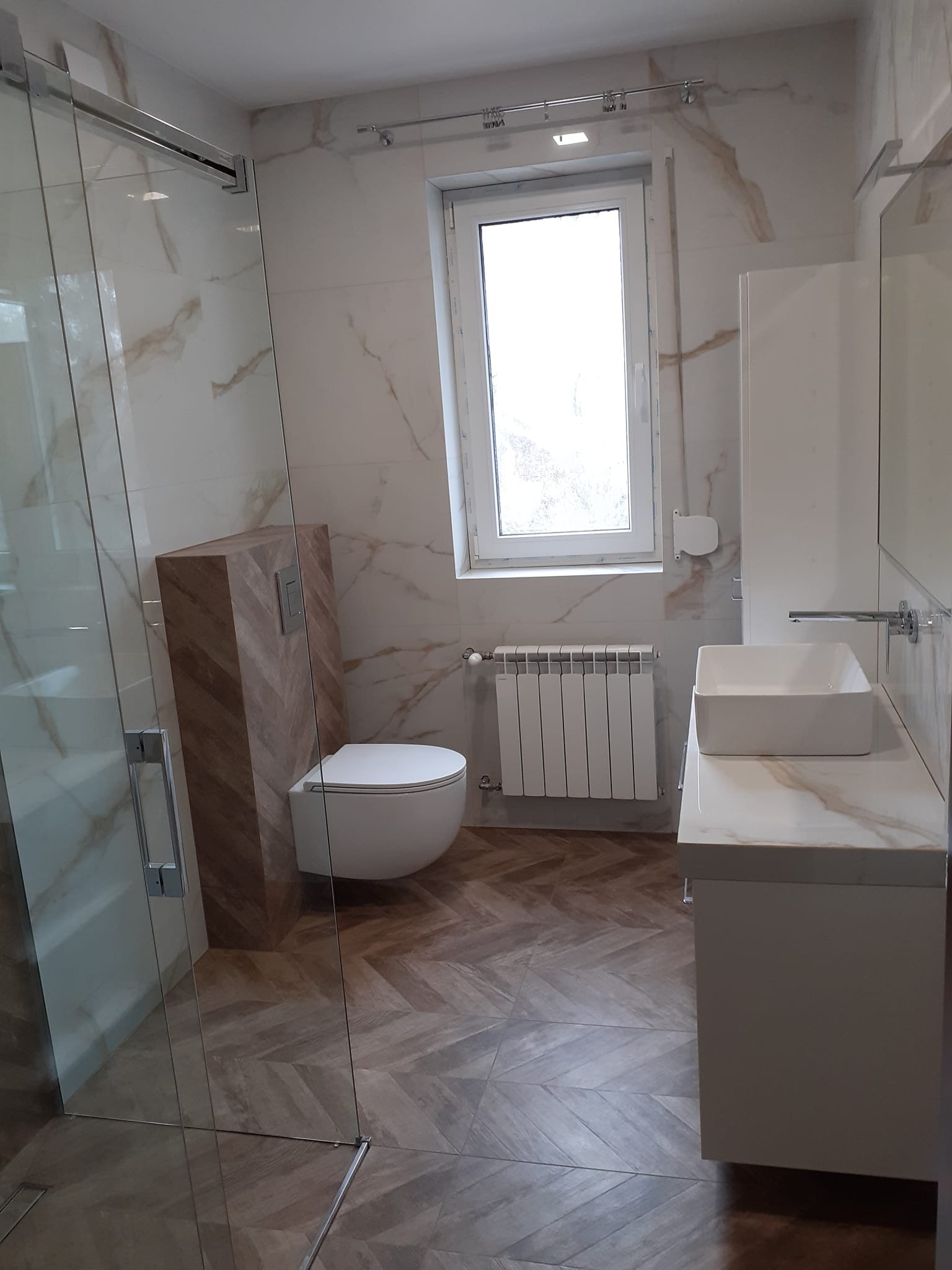 Elegancka łazienka po remoncie z kabiną prysznicową, toaletą w zabudowie z drewnianą mozaiką i umywalką na blacie z marmurowym wzorem. Parkiet ułożony w jodełkę.