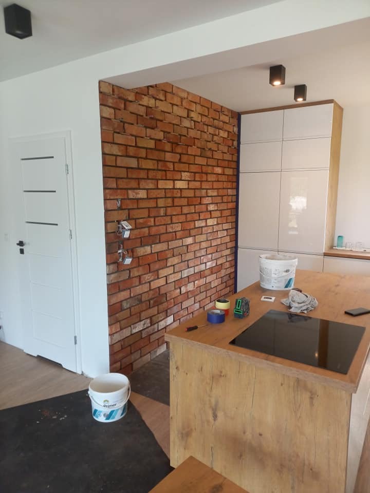 Ceglana ściana w salonie w trakcie wykańczania, widoczne narzędzia i materiały budowlane. Elementy kuchni w tle, białe ściany i drzwi.