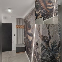 BIPI-BUD Bartłomiej Pazdro - Przedpokój z fototapetą przedstawiającą twarz kobiety w stylu vintage, oświetlony pionową listwą LED, szara tapicerowana ściana z wieszakami i siedziskiem, szara podłoga z płytek.