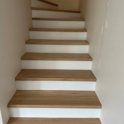 BIPI-BUD Bartłomiej Pazdro - Jasne, drewniane schody z białymi podstopnicami, widziane z dołu. Naturalne oświetlenie podkreśla strukturę drewna i minimalistyczny design. Ściany w odcieniach beżu.