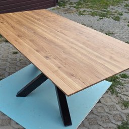 BIPI-BUD Bartłomiej Pazdro - Prostokątny blat drewniany z wyraźnym usłojeniem, wsparty na czarnych, metalowych nogach w kształcie litery X. Ustawiony na niebieskiej płycie na zewnątrz.