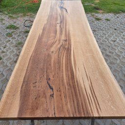 BIPI-BUD Bartłomiej Pazdro - Długi, gotowy blat z litego drewna dębowego z wyraźnym usłojeniem i żywicą epoksydową w naturalnym oświetleniu zewnętrznym, na tle kostki brukowej i trawy.