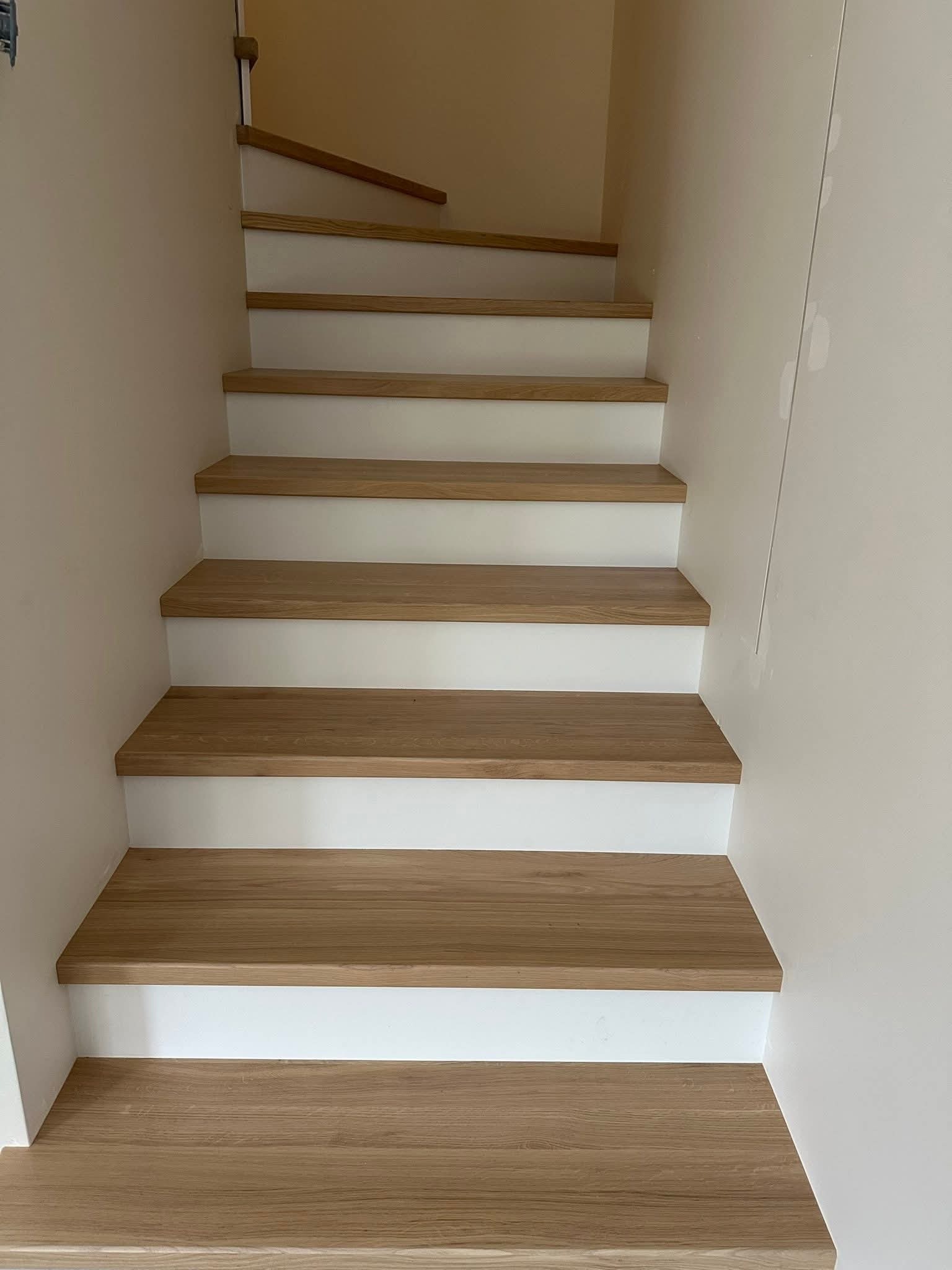 Jasne, drewniane schody z białymi podstopnicami, widziane z dołu. Naturalne oświetlenie podkreśla strukturę drewna i minimalistyczny design. Ściany w odcieniach beżu.