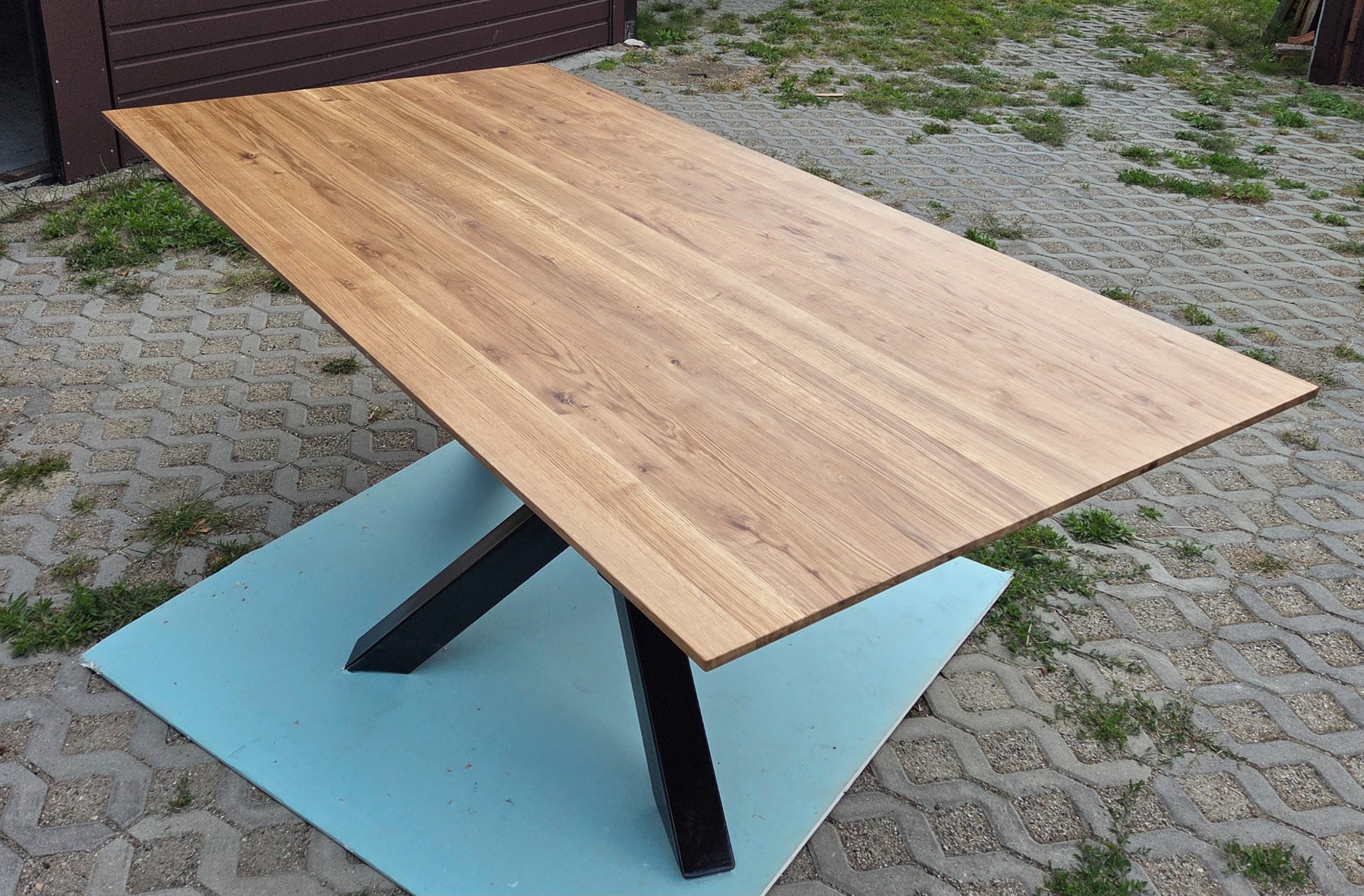Prostokątny blat drewniany z wyraźnym usłojeniem, wsparty na czarnych, metalowych nogach w kształcie litery X. Ustawiony na niebieskiej płycie na zewnątrz.