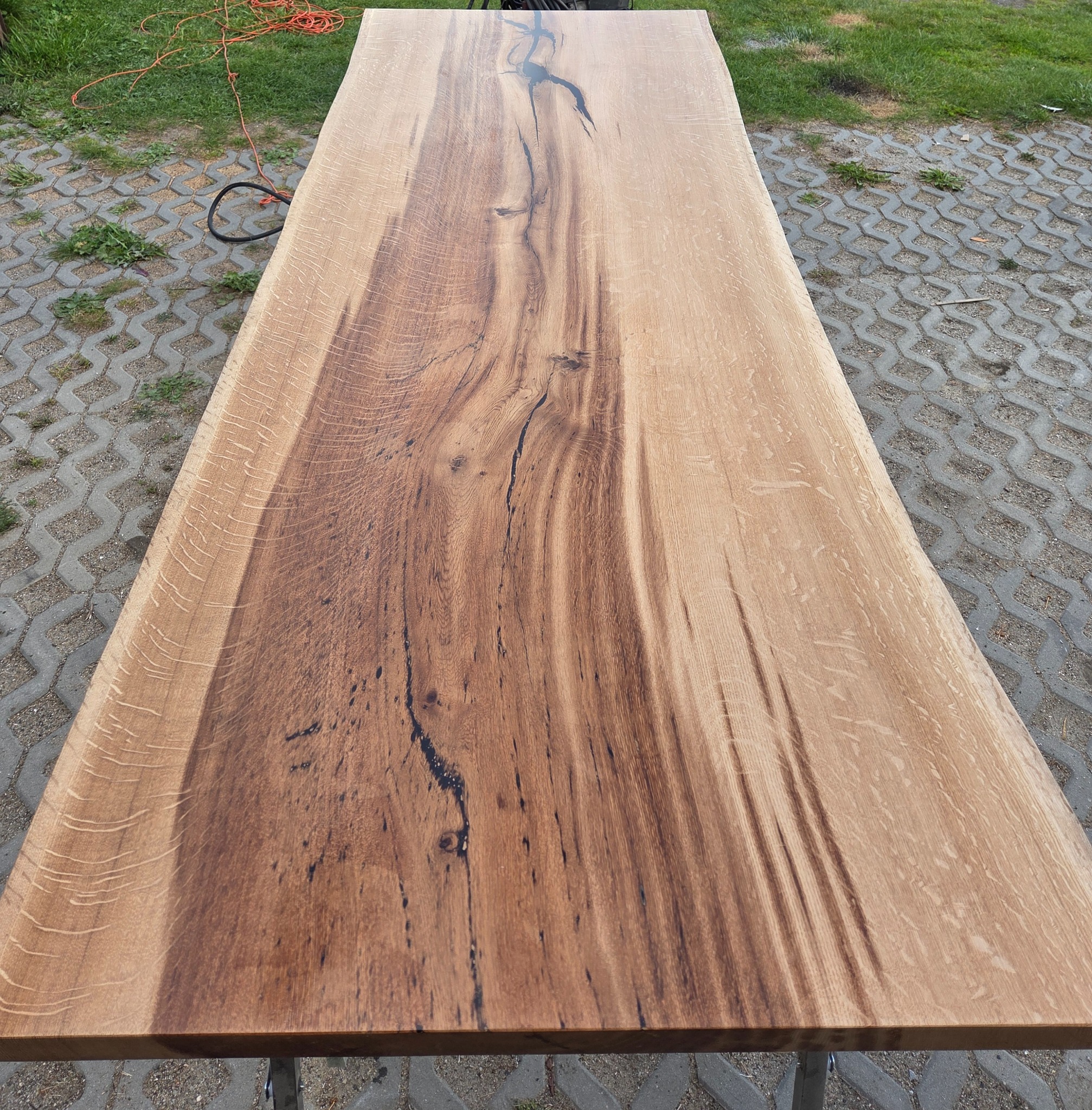 Długi, gotowy blat z litego drewna dębowego z wyraźnym usłojeniem i żywicą epoksydową w naturalnym oświetleniu zewnętrznym, na tle kostki brukowej i trawy.