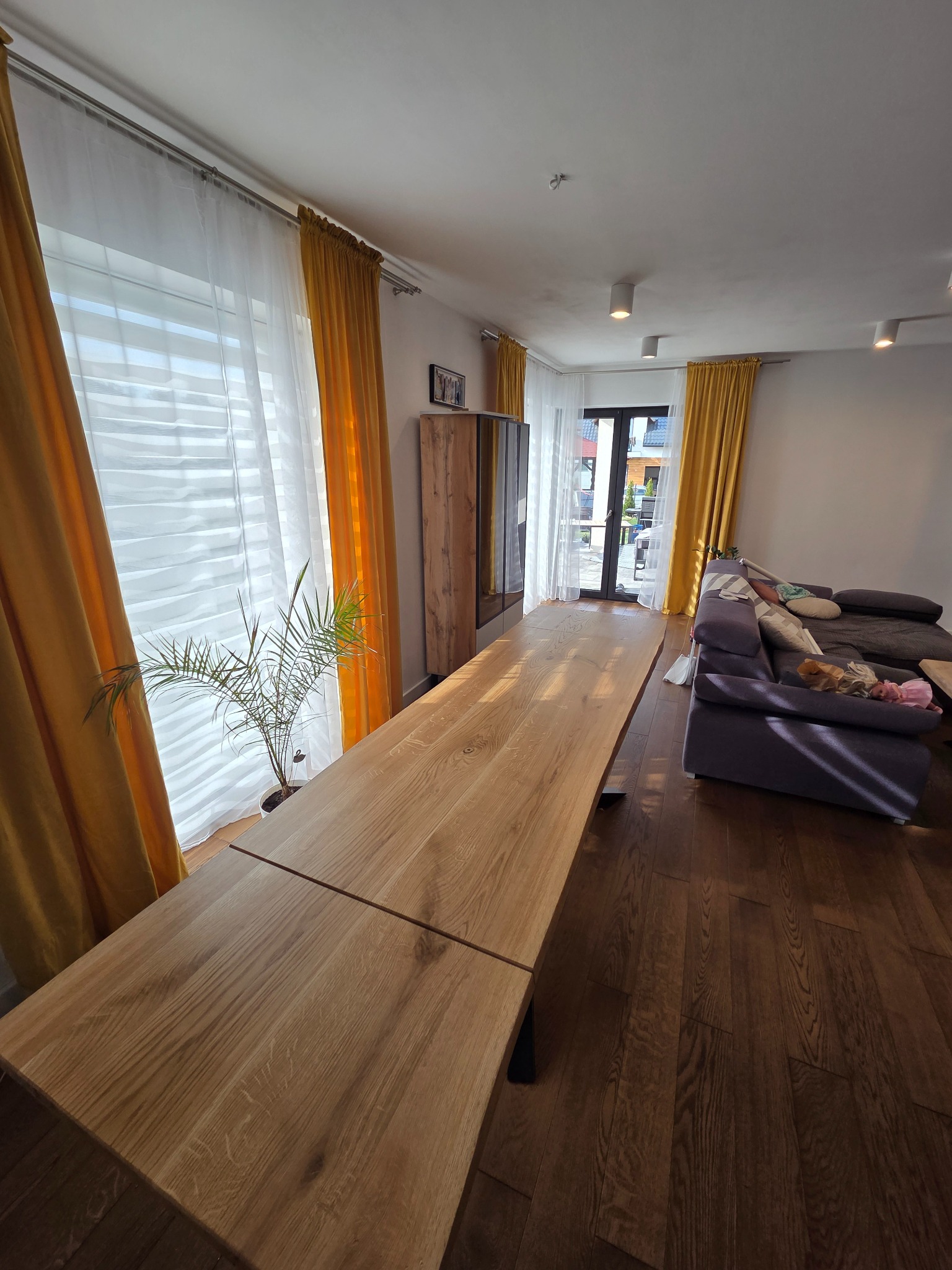 Rozłożony, drewniany stół w salonie z parkietem i żółtymi zasłonami. Stół z blatem z wyraźnym usłojeniem, ustawiony w jasnym wnętrzu.