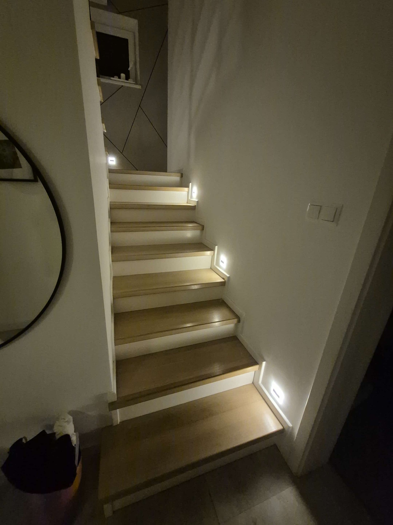 Drewniane schody z podświetleniem LED wzdłuż stopni, widok z dołu. Minimalistyczny design, jasne drewno, białe ściany. Okrągłe lustro w tle.