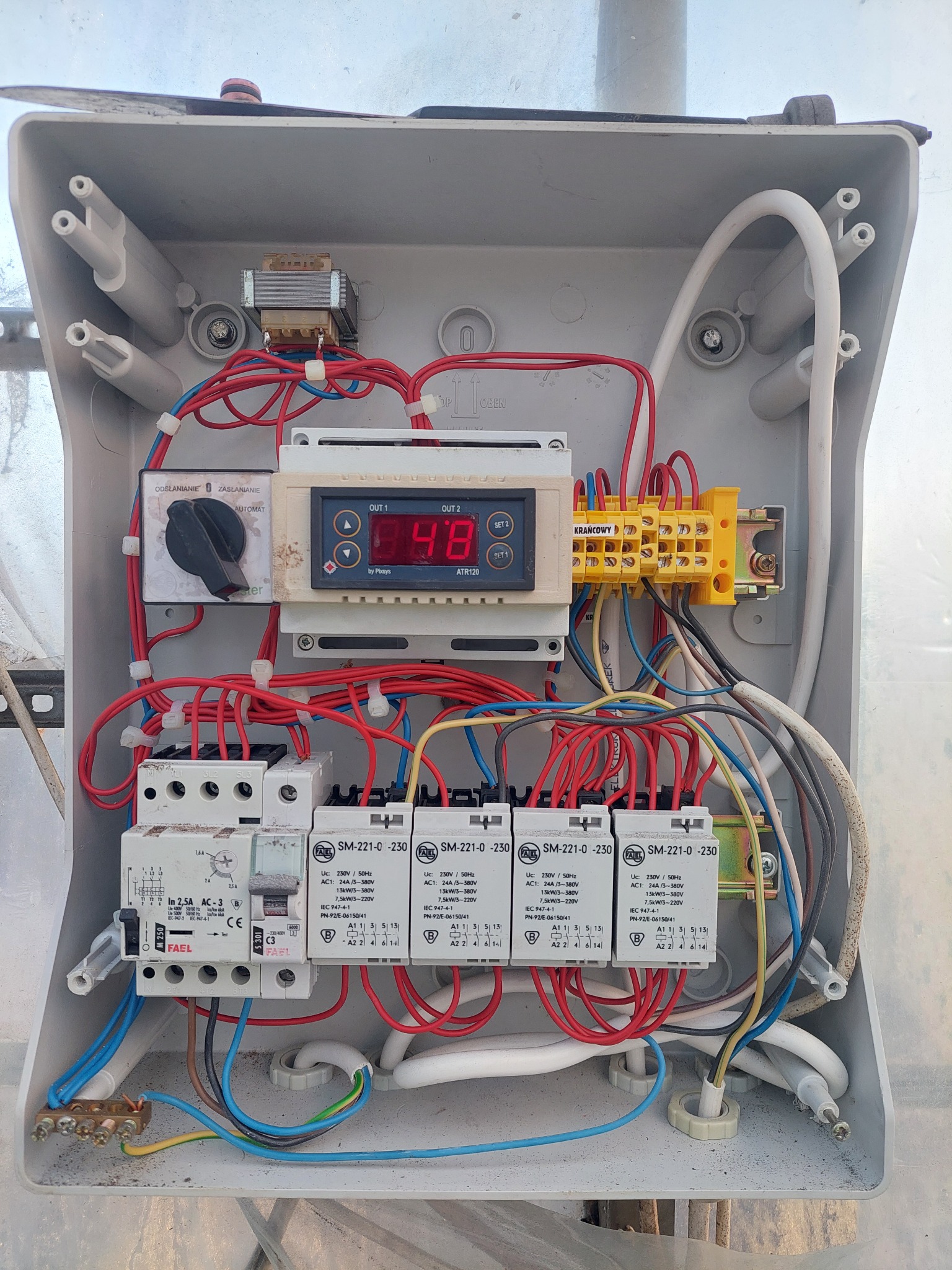Otwarta skrzynka elektryczna z widocznymi elementami sterującymi, transformatorem, przekaźnikami FAEL, zabezpieczeniem i licznymi przewodami w różnych kolorach, zamontowana na zewnątrz.