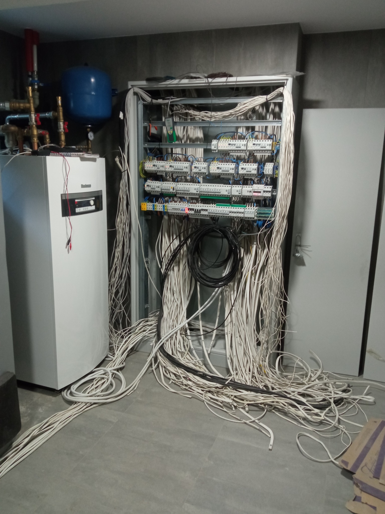 Rozdzielnica elektryczna z licznymi przewodami w trakcie montażu w Kaliszu, widoczny również piec CO i zbiornik wyrównawczy.