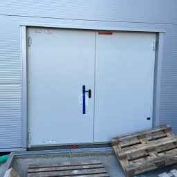 AZ SERVIS SPÓŁKA Z OGRANICZONĄ ODPOWIEDZIALNOŚCIĄ - Metalowe dwuskrzydłowe drzwi techniczne w szarej fasadzie budynku przemysłowego. Palety drewniane leżą na zewnątrz, przed wejściem.
