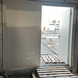 AZ SERVIS SPÓŁKA Z OGRANICZONĄ ODPOWIEDZIALNOŚCIĄ - Częściowo otwarta, biała brama przemysłowa z widokiem na plac budowy. Palety drewniane na podłodze wewnątrz budynku. Surowy betonowy strop.