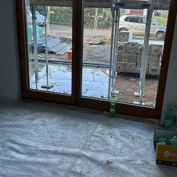 AZ SERVIS SPÓŁKA Z OGRANICZONĄ ODPOWIEDZIALNOŚCIĄ - Montaż okien balkonowych z brązową ramą w surowym stanie. Na zewnątrz widoczne rusztowania, materiały budowlane i dom.