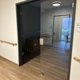 AZ SERVIS SPÓŁKA Z OGRANICZONĄ ODPOWIEDZIALNOŚCIĄ - Szklane drzwi wewnętrzne z czarną ramą, widok na korytarz z drewnianą podłogą i poręczą. W tle paczki i widok na ogród przez kolejne drzwi.