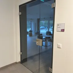 Szklane drzwi z metalową ramą, prowadzące do kawiarni z widocznym wnętrzem i oznaczeniem numeru pokoju 0.03, oświetlone światłem dziennym.