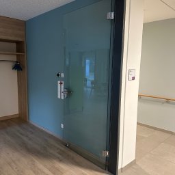 AZ SERVIS SPÓŁKA Z OGRANICZONĄ ODPOWIEDZIALNOŚCIĄ - Szklane drzwi z czarną ramą, zamek elektroniczny, widok na wnętrze. Podłoga z paneli, błękitna ściana. Widoczna klamka i zawiasy. Minimalistyczny design.