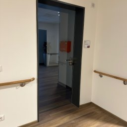 AZ SERVIS SPÓŁKA Z OGRANICZONĄ ODPOWIEDZIALNOŚCIĄ - Szklane drzwi wewnętrzne z czarną ramą w jasnym pomieszczeniu, widoczna apteczka pierwszej pomocy w tle. Podłoga z paneli imitujących drewno.