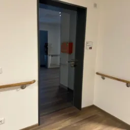 Szklane drzwi wewnętrzne z czarną ramą w jasnym pomieszczeniu, widoczna apteczka pierwszej pomocy w tle. Podłoga z paneli imitujących drewno.