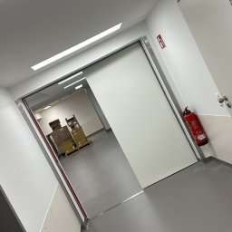 AZ SERVIS SPÓŁKA Z OGRANICZONĄ ODPOWIEDZIALNOŚCIĄ - Nowoczesne, przesuwne drzwi wewnętrzne z czerwonym znakiem ewakuacyjnym i gaśnicą w tle, w sterylnym, białym korytarzu, ujęcie pod kątem.