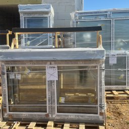 AZ SERVIS SPÓŁKA Z OGRANICZONĄ ODPOWIEDZIALNOŚCIĄ - Nowe okna PCV Salamander, zabezpieczone folią, ułożone na paletach na placu budowy. Widoczne cegły i narzędzia. Słoneczny dzień.