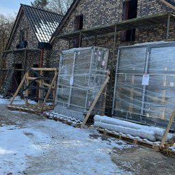 AZ SERVIS SPÓŁKA Z OGRANICZONĄ ODPOWIEDZIALNOŚCIĄ - Nowe okna PCV w folii na paletach, czekają na montaż przy domu z cegły w zimowej scenerii. Widoczne rusztowania i przygotowania do instalacji.
