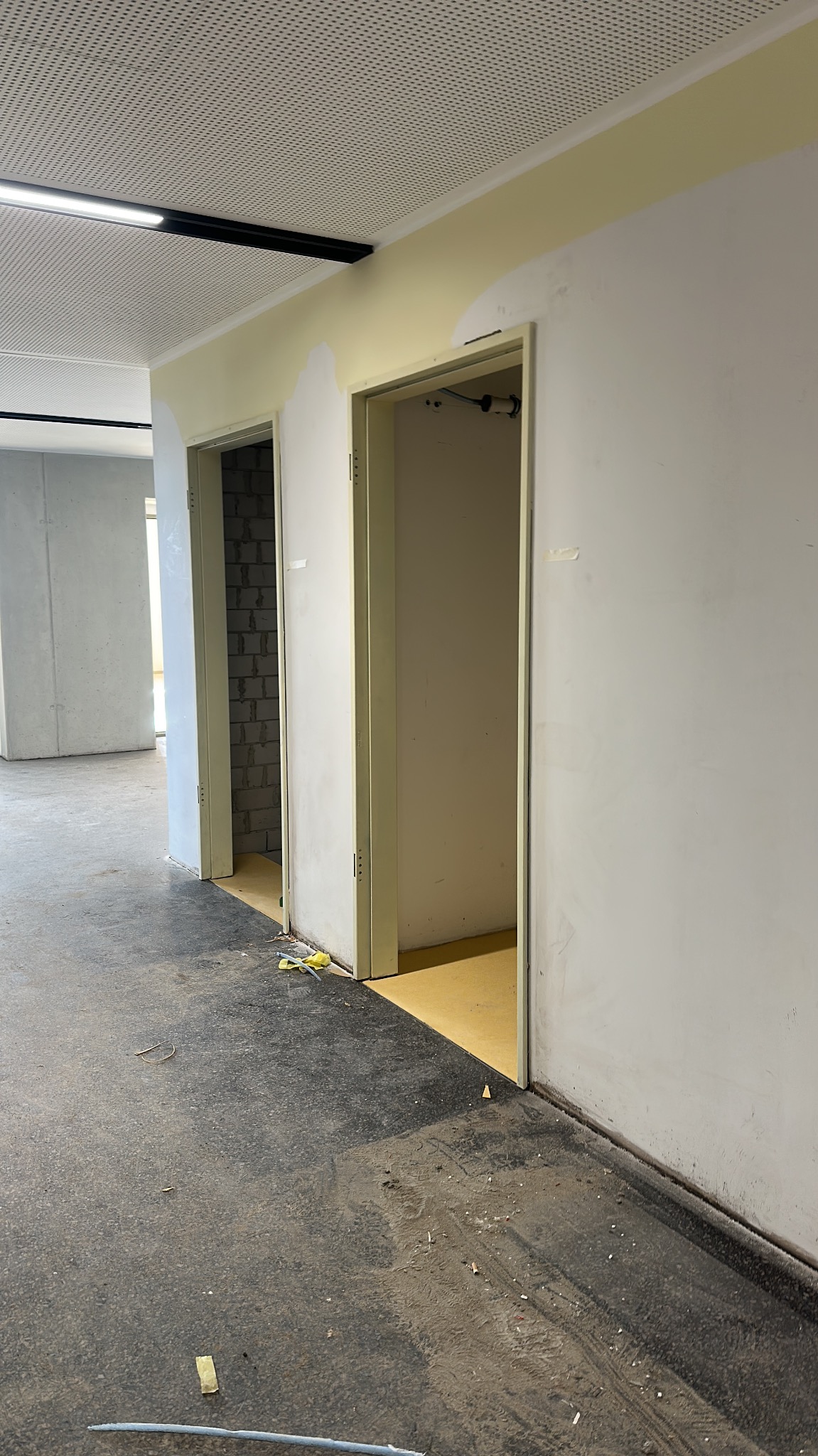 Wnętrze w trakcie remontu: widoczne dwa otwarte, białe ościeża drzwiowe, świeżo pomalowane ściany w odcieniach bieli i żółci, oraz ciemna podłoga z zabrudzeniami budowlanymi.