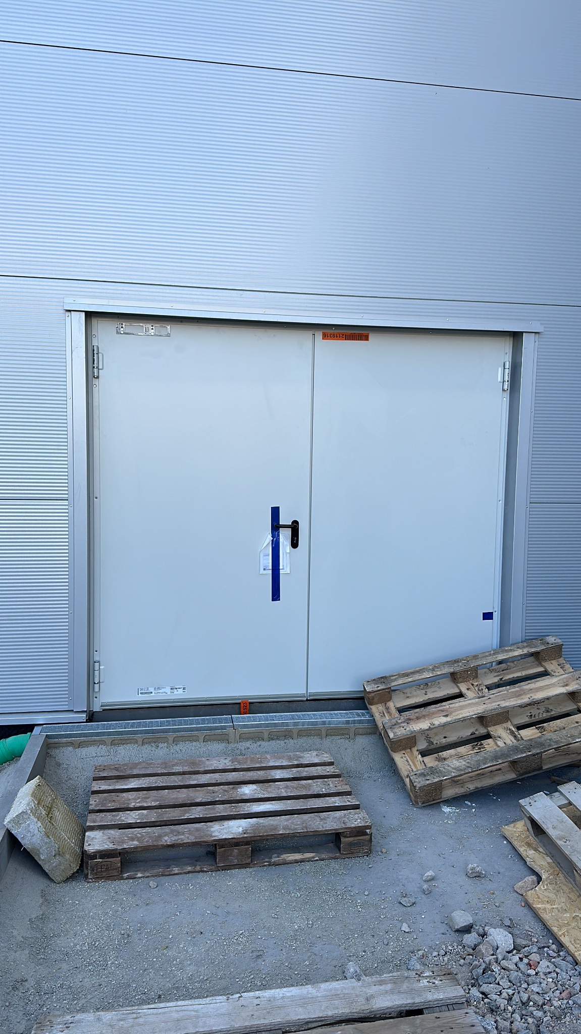 Metalowe dwuskrzydłowe drzwi techniczne w szarej fasadzie budynku przemysłowego. Palety drewniane leżą na zewnątrz, przed wejściem.