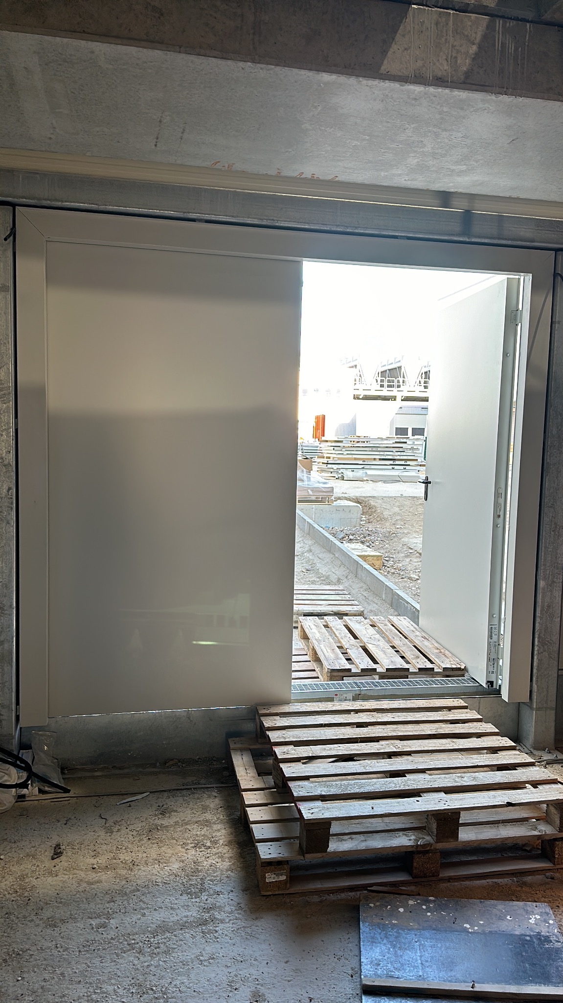 Częściowo otwarta, biała brama przemysłowa z widokiem na plac budowy. Palety drewniane na podłodze wewnątrz budynku. Surowy betonowy strop.