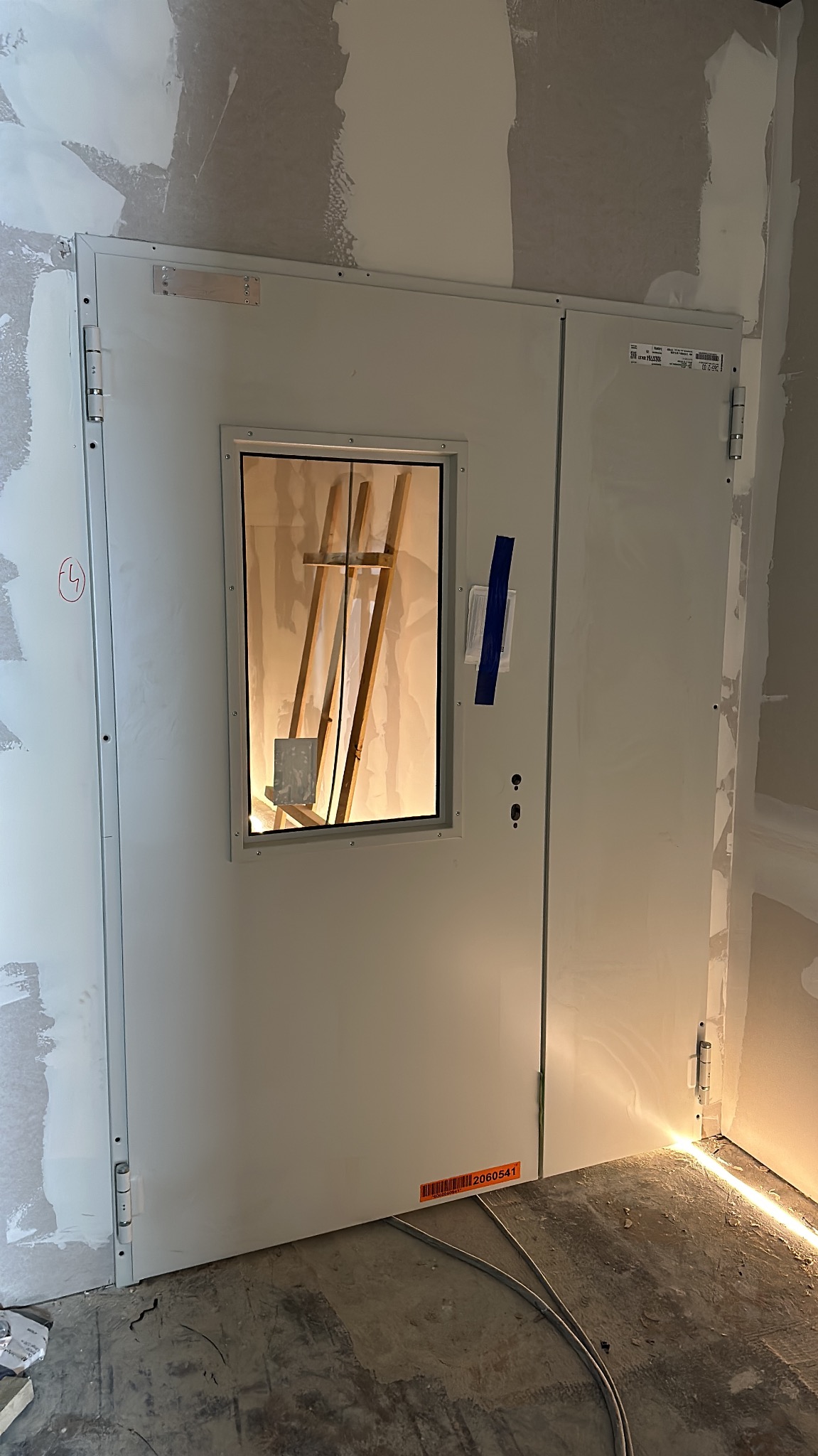 Białe metalowe drzwi techniczne z małym okienkiem i niebieską taśmą na tle surowej ściany w trakcie remontu. Widoczne kable na podłodze i elementy rusztowania wewnątrz.