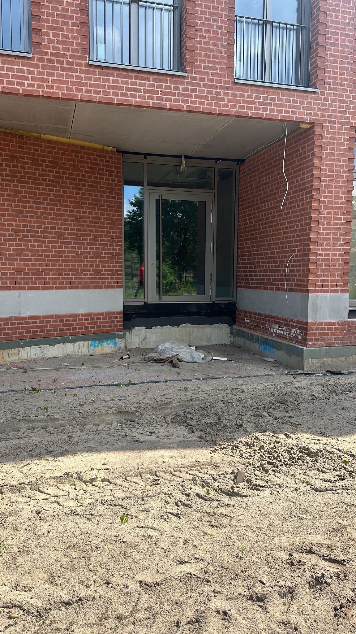 Nowe, szklane drzwi wejściowe w budynku z czerwonej cegły, widoczne przewody i piasek przed wejściem. Etap prac wykończeniowych.