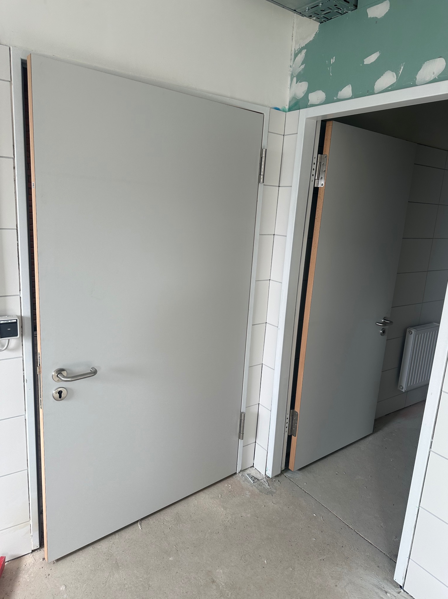 Dwa szare skrzydła drzwiowe z klamkami, zamontowane w białej ościeżnicy. W tle widoczne białe płytki i grzejnik, surowy strop i fragment ściany po szpachlowaniu.